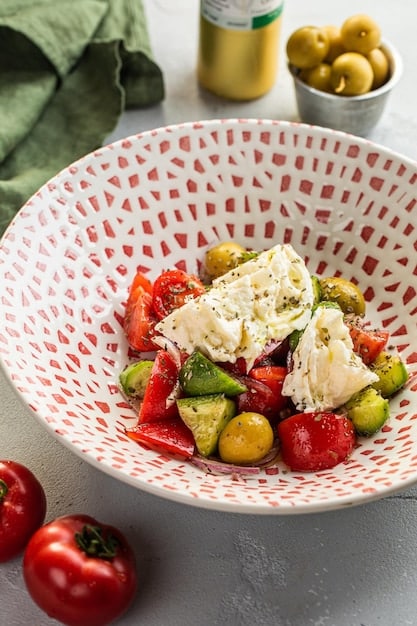 Close-up shot of a vibrant Greek salad with chunks of feta cheese, ripe tomatoes, cucumber slices, Kalamata olives, and red onion rings, all tossed in a glistening olive oil and oregano dressing. Grilled chicken strips are artfully arranged on top.