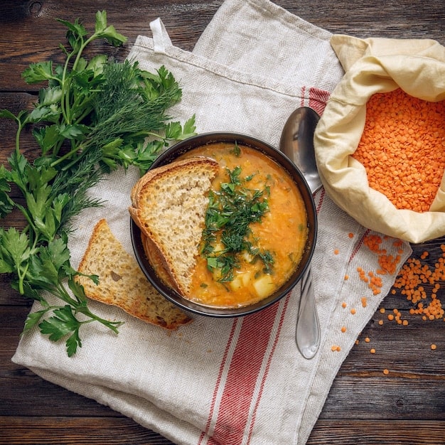 A bowl of hearty lentil soup, garnished with a sprig of fresh parsley and a drizzle of olive oil. A slice of whole-grain bread sits beside the bowl, with its texture clearly visible. The background is a warm, rustic kitchen setting.
