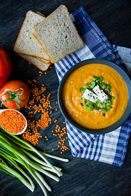 A top-down shot of a bowl of lentil soup with a slice of whole-grain bread on the side, garnished with fresh parsley. The soup is colorful and textured, showcasing various vegetables and lentils.