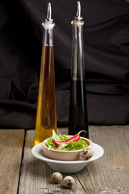 A small glass bottle filled with homemade balsamic vinaigrette. The vinaigrette is dark brown and has visible herbs floating inside. The bottle is placed on a rustic wooden table next to a bowl of mixed green salad, drizzled with the same vinaigrette. A small dish of balsamic vinegar and a bottle of olive oil are also present, contributing to the scene.