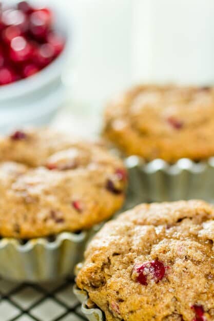 An eye-level shot of three cranberry muffins on a cooling rack, with visible cranberries throughout and a light dusting of powdered sugar on top, emphasizing the colorful and festive appearance of the treats.