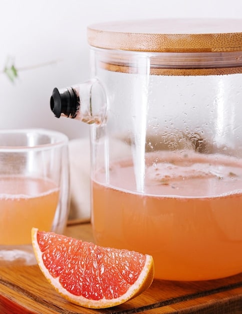 A brightly lit image of a glass dispenser filled with grapefruit and ginger detox water. Thin slices of grapefruit and ginger are visible, floating throughout the water. The dispenser is set against a clean, modern background.