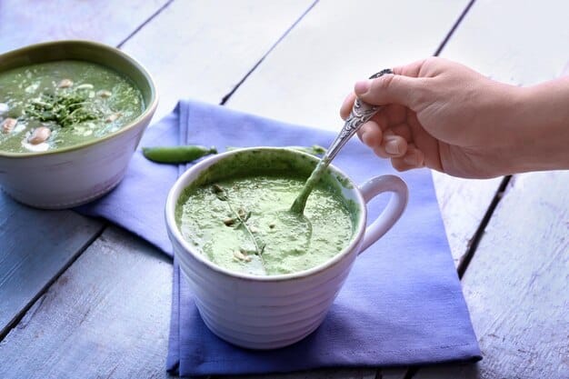 A close-up shot of a creamy green detox soup being poured into a white ceramic bowl. The soup has visible chunks of blended vegetables and is topped with a swirl of coconut milk and a sprinkle of chia seeds.
