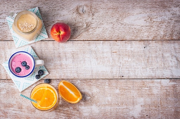 An overhead shot of a wooden table with an array of smoothie ingredients, including oranges, kiwis, pineapples, spinach, and a bottle of almond milk. Next to it is a blender filled halfway with a blended smoothie.