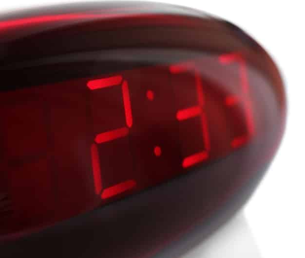A close-up of a digital timer displaying the time 1:30 PM, with a blurred background of a kitchen counter where fruits, nuts, and a blender are visible. This emphasizes the importance of timing when preparing and consuming pre-workout meals.